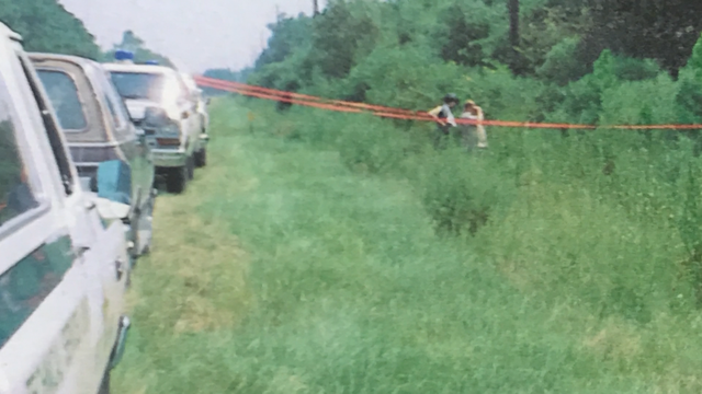 A crime scene photograph showing a rural road bordered by thick Florida brush, pine trees, and overgrown foliage. Yellow police line tape is strung across the frame, blocking access to the wooded area off the side of the road.