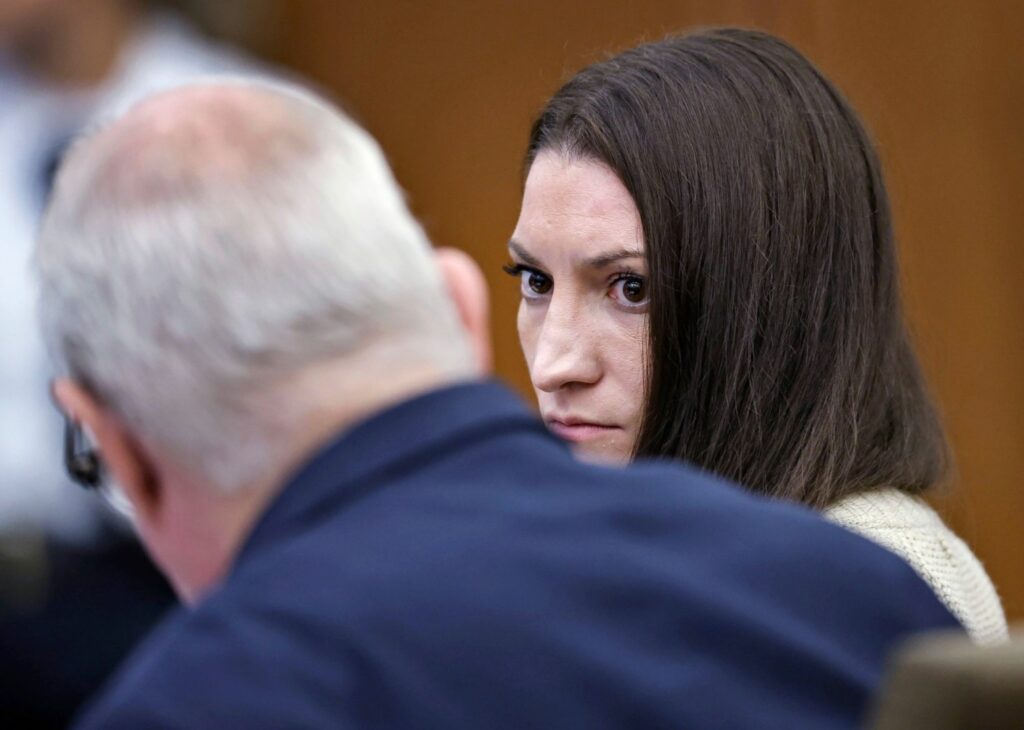 Lindsay Clancy in courtroom talking to her lawyer with a focused look on her face with long dark hair and a beige sweater on.