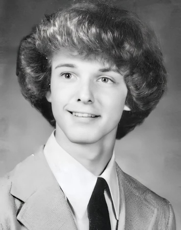 Black and white high school yearbook portrait of a young Pamela Leigh Walton, pictured prior to her transition as a clean-shaven male youth with short, neatly combed dark hair, wearing a dark formal suit jacket and white collared shirt.
