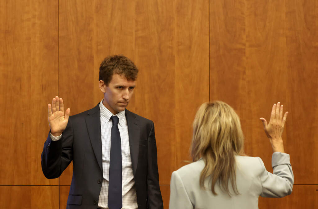 patrick Clacy in the courtroom being sworn in with his right hand raised wearing a blue suit and tie.