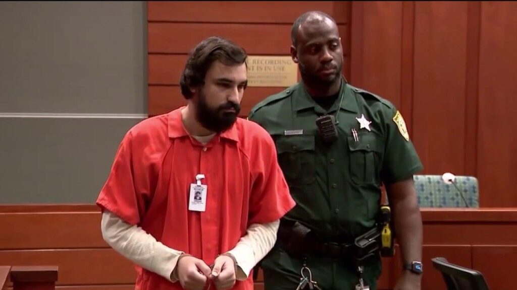 Ahmad Jihad Bojeh stands in a Florida courtroom next to a police officer during a legal proceeding regarding his mental evaluation.