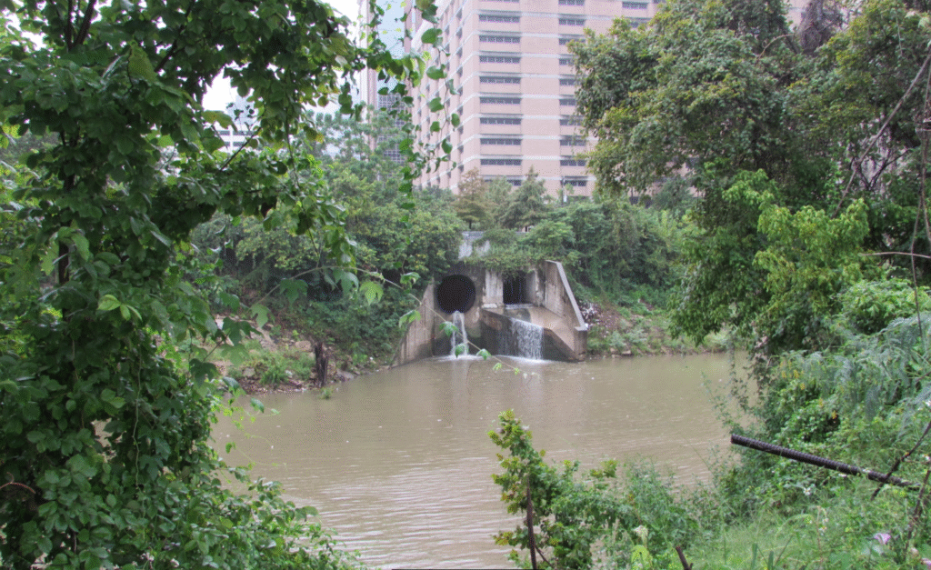 A daytime view of a concrete storm drain outlet pouring rushing water directly into the wide, muddy channel of Buffalo Bayou with the downtown Houston urban landscape nearby.