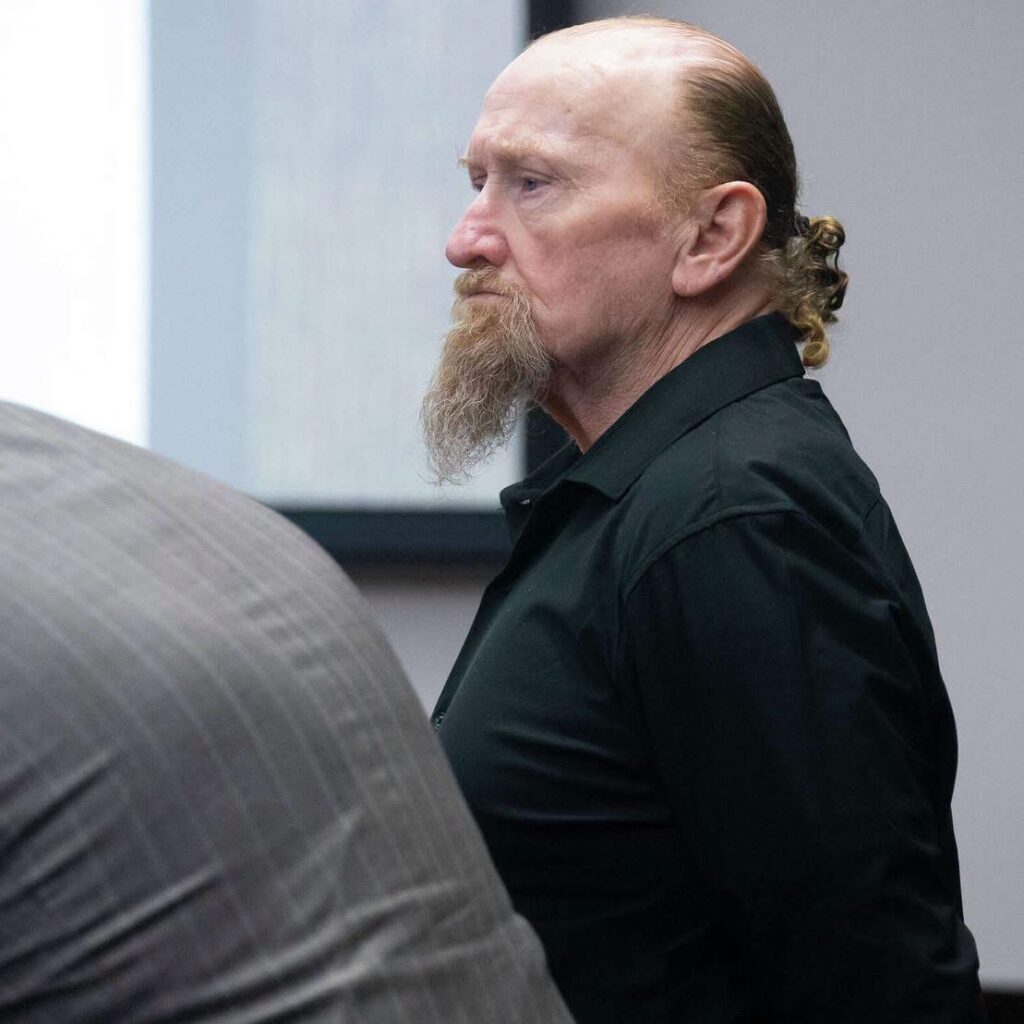 Old red hair man with pony tail in a black suit standing in a courtroom.