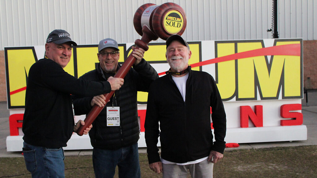 Three smiling men posing together outdoors while holding a giant wooden novelty courtroom mallet.