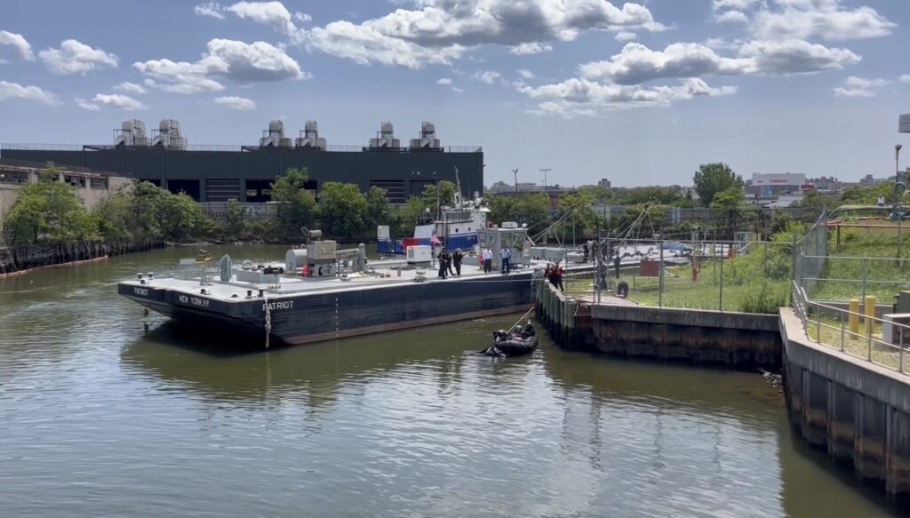 An NYPD harbor unit boat on Newtown Creek during the day, with officers actively recovering a body from the waterway.
