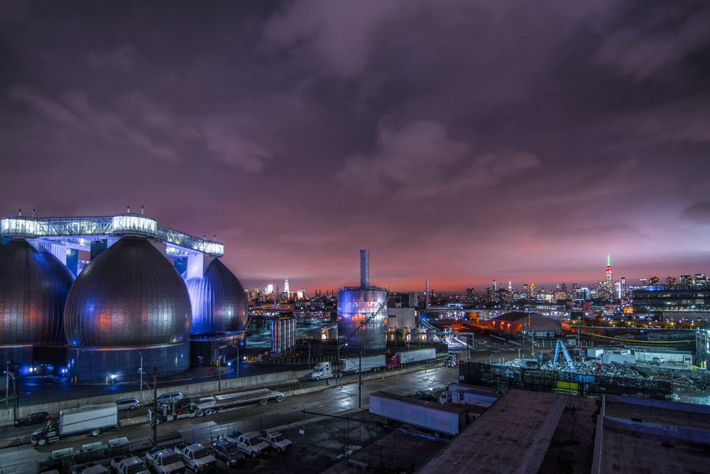A stark night view of a completely empty, industrial street leading toward the Newtown Creek wastewater facility, where massive, egg-shaped metallic structures glow with light against the dark sky.