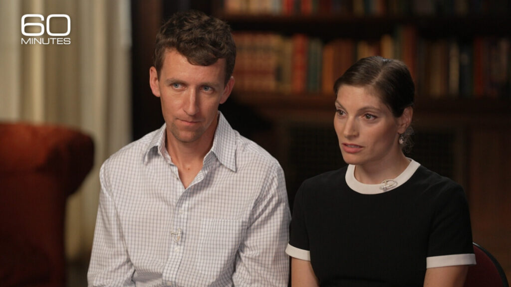 Patrick Clancy and his wife, Dr. Rachel Danis, sitting close together during their joint appearance on CBS News' 60 Minutes. Both are dressed in casual, respectful attire, looking towards the interviewer with supportive and serious expressions.