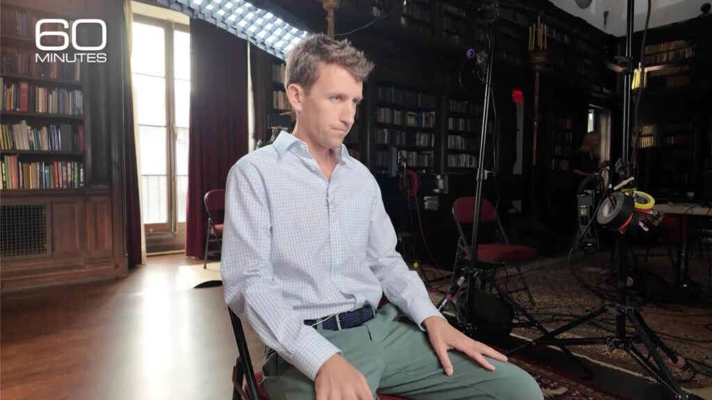 A medium shot of Patrick Clancy sitting in a chair during a formal television interview environment. He is dressed in a dark suit jacket and looking slightly off-camera with a somber expression. Professional studio lighting and a blurred background frame the emotional interview setting.