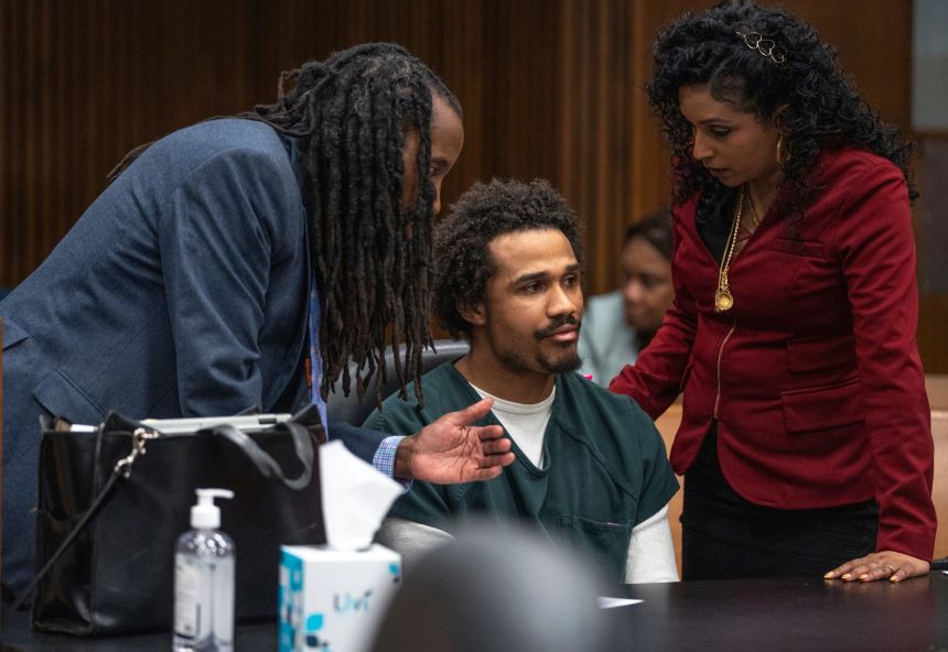 A courtroom photograph of Michael Manuel Jackson-Bolanos seated and wearing a forest green prison uniform. He is leaning slightly to speak or listen attentively to his defense attorneys who are sitting beside him. The background shows the professional setting of a courtroom.