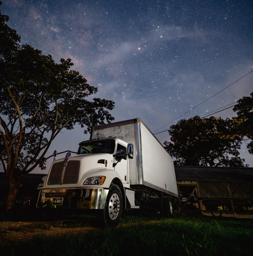 A lone semi-truck trailer parked on a dark, isolated asphalt lot at night, illuminated only by a distant, dim street lamp under a pitch-black sky.