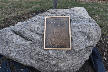 A bronze memorial plaque with the names and etched portraits of Andrew, Alexander, and Tanner Skelton attached to a large decorative rock outdoors, with the words "Faith, Hope, Love" visible.