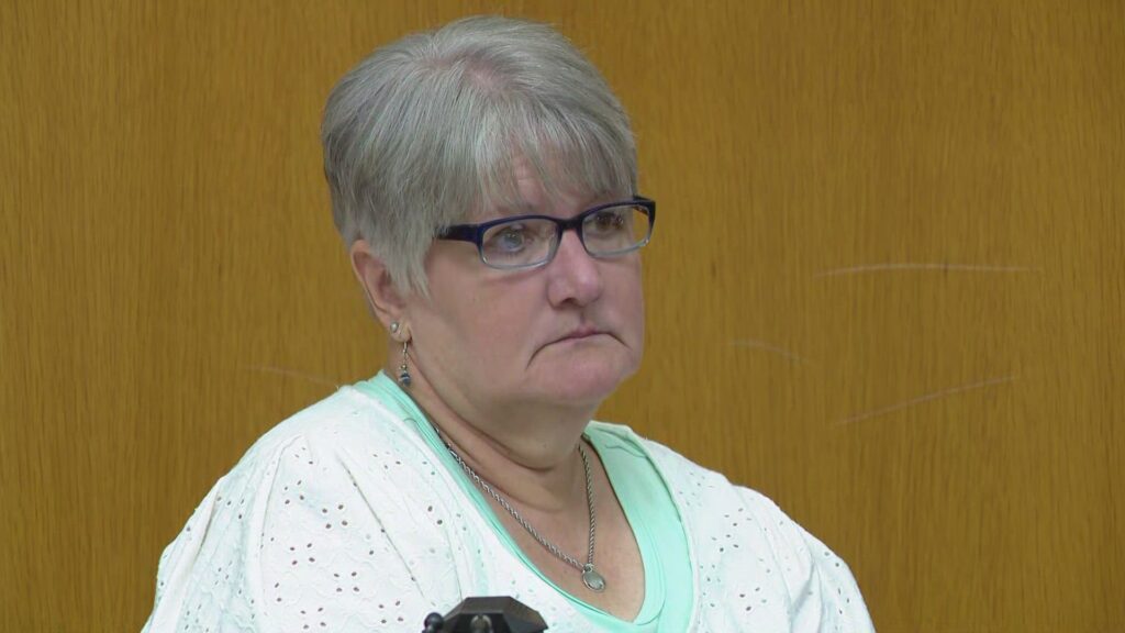 A woman in a courtroom setting, seated near a wooden partition, wearing a white sweater and mint green top with black glasses with a serious expression on her face