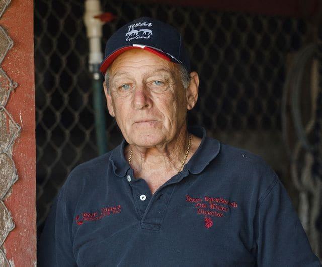 Older man standing in a barn or stable wearing a blue polo shirt AND BASEBALL CAP