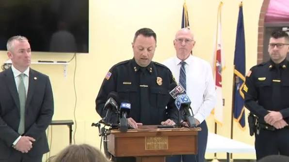 Mechanicville Police Chief William Rabbitt speaks from a podium during an official law enforcement press conference. He is wearing his uniform, standing in front of microphones with official seals in the background.