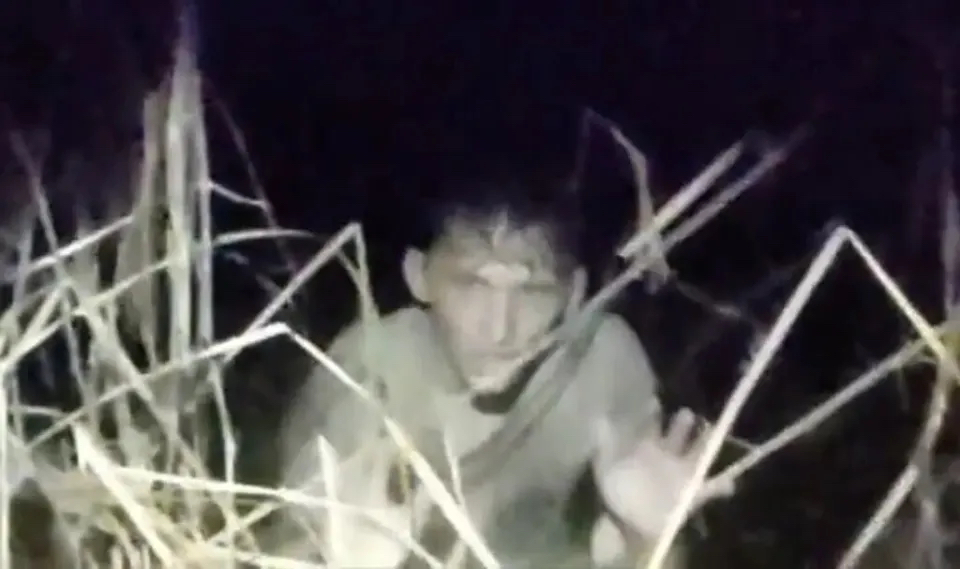A police bodycam still showing Isaac Kinsey emerging from thick swamp weeds and murky water with his hands up in the air.
